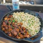 A cast iron skillet filled with BBQ chicken and beer cheese macaroni and a can of Samuel Adams in the background
