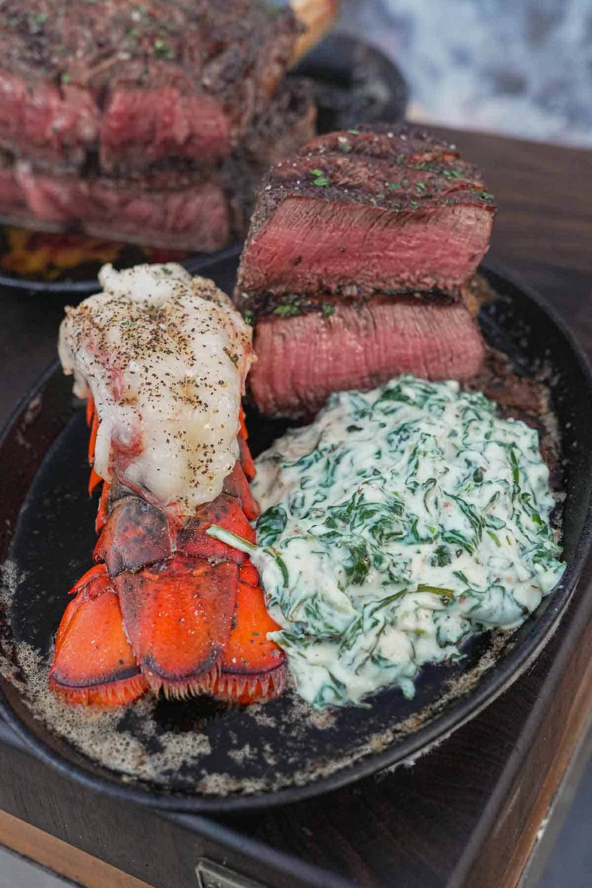 A cooked lobster tail, steaks cut open to show the inside and creamed spinach on a plate