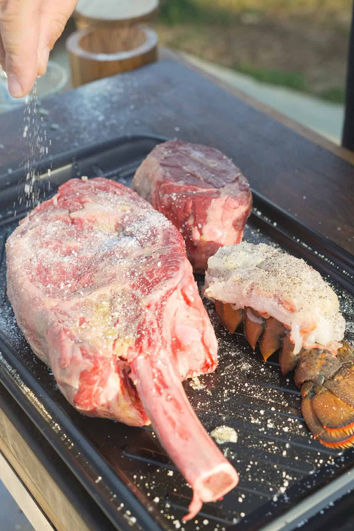 Seasoning bone-in ribeye steak and filet and lobster tail with kosher salt