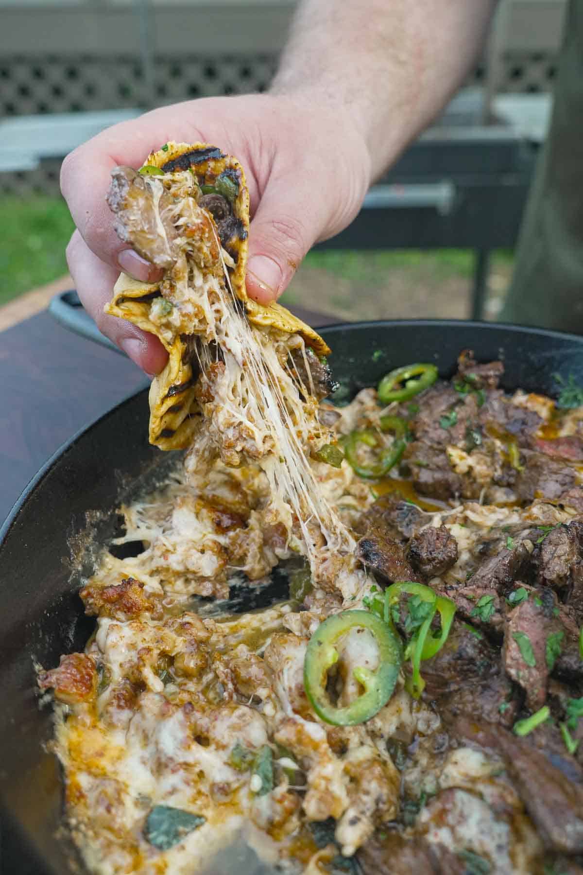 A hand using a tortilla to pull queso flameado out of a cast iron skillet