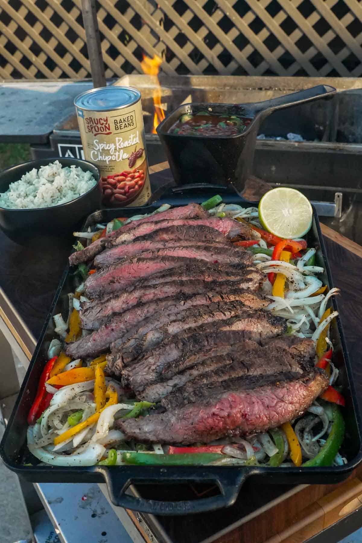 Plated steak fajitas with bell peppers, onions, sliced limes, rice and Saucy Spoon baked beans in the background