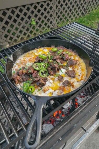 A cast iron skillet of queso flameado on the grill, with chopped cilantro and jalapeño slices for the garnish