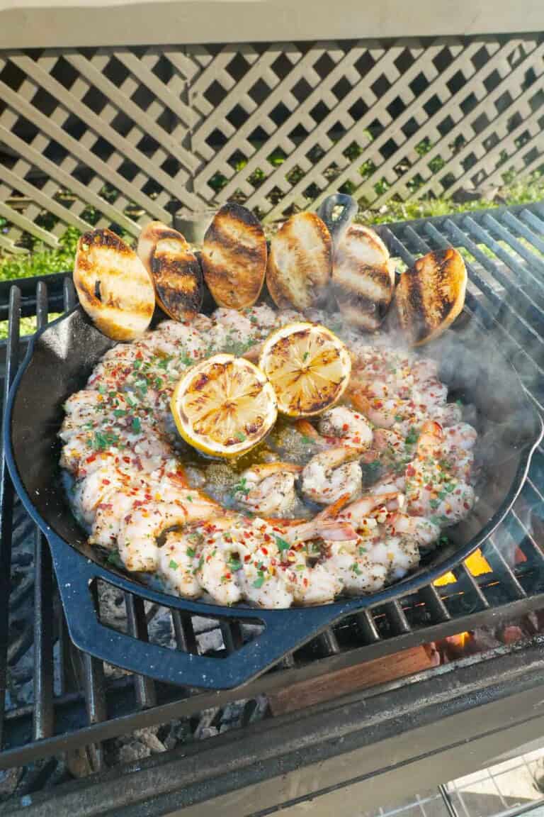 A skillet on the grill filled with smoked shrimp, charred lemon, chopped fresh parsley and grilled bread slices