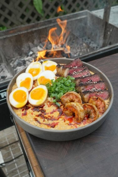 A full bowl of surf and turf ramen next to the grill, filled with steak, shrimp, eggs, noodles and garnishes