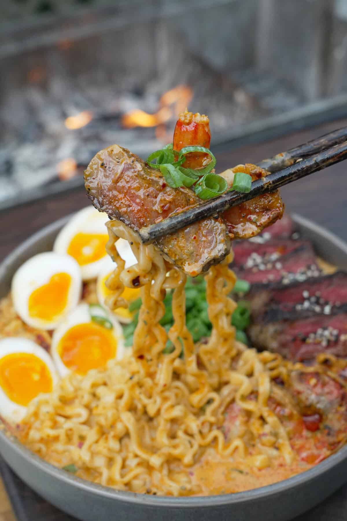 Chopsticks holding up a piece of steak from a bowl of surf and turf ramen with eggs, shrimp, noodles and scallions