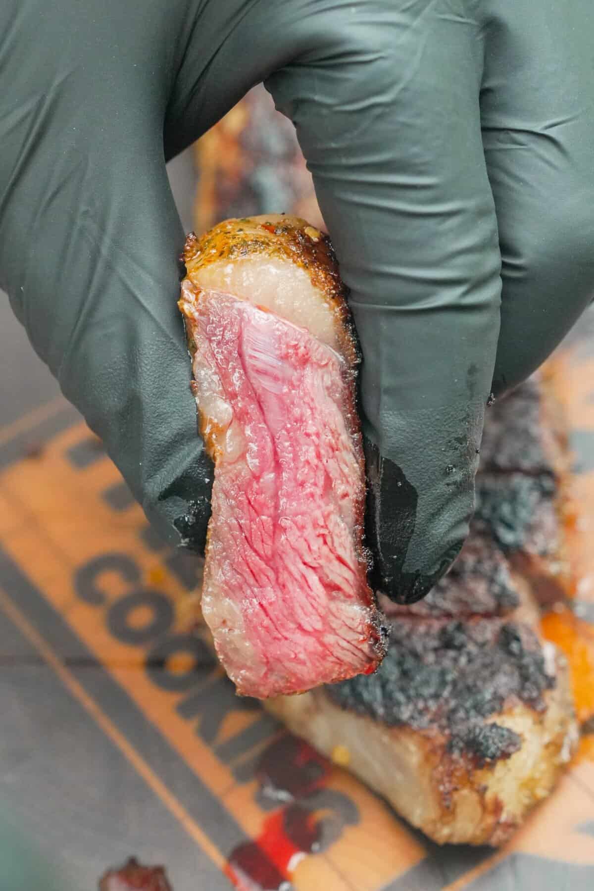 A gloved hand holding a slice of grilled New York strip steak