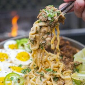 A hand holding chopsticks and pulling up noodles of birria ramen, which is garnished with soft boiled eggs, chopped cilantro and diced red onion
