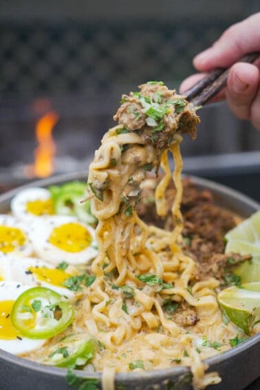 A hand holding chopsticks and pulling up noodles of birria ramen, which is garnished with soft boiled eggs, chopped cilantro and diced red onion