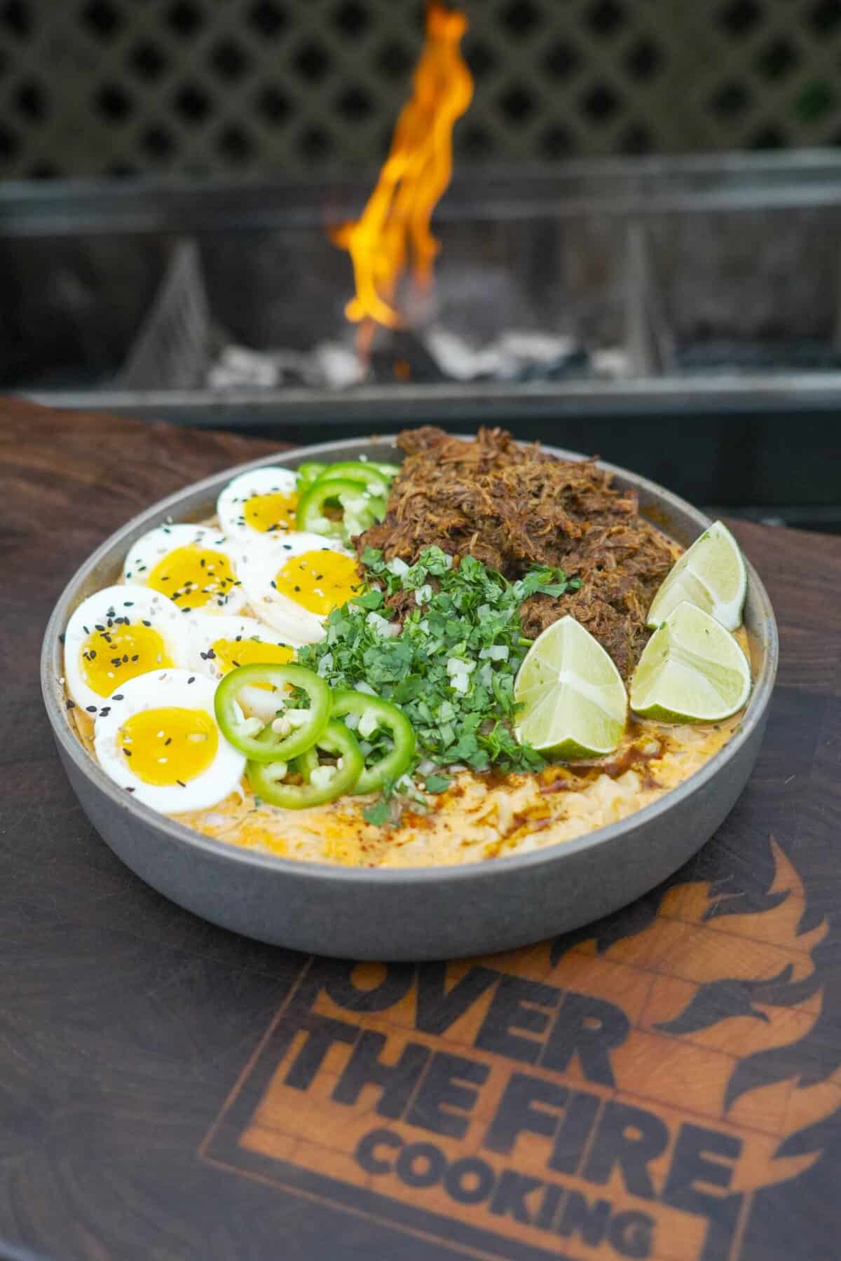 A bolw of birria ramen with beef, eggs, noodles and garnishes in front of the grill