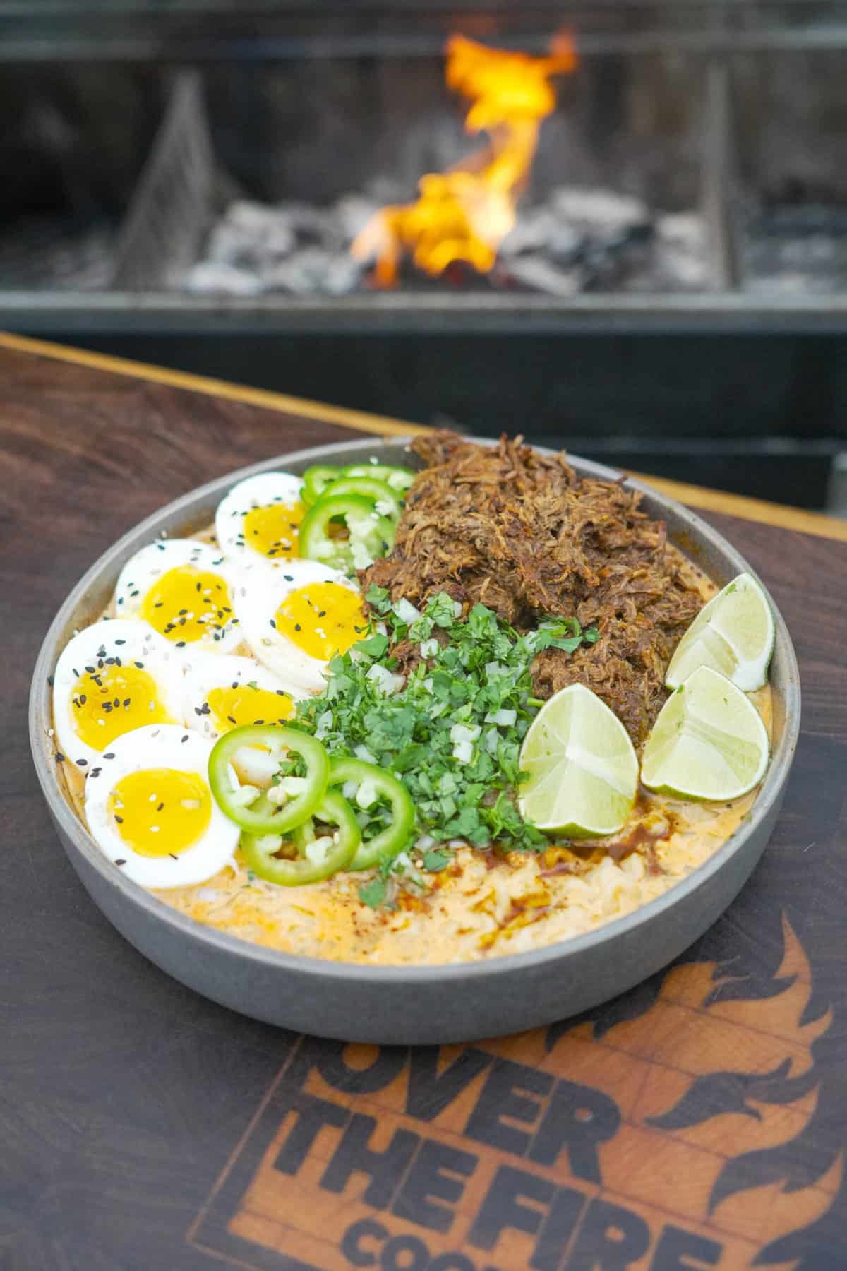 A bowl of birria ramen with all the fixings, placed in front of a flame on the grill