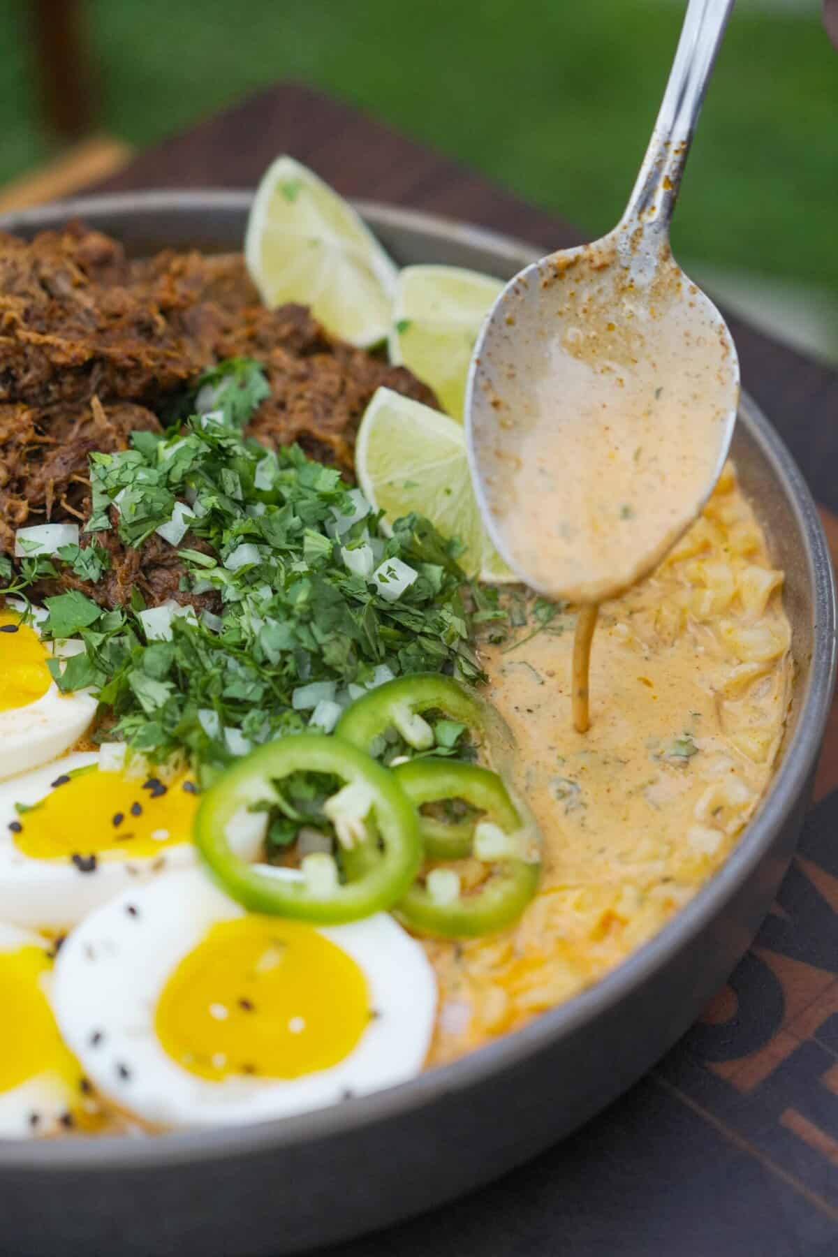 A spoonful of ramen broth from a bowl of birria ramen with sliced jalapeńos, cilantro, eggs and beef