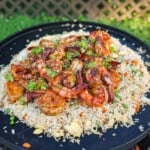 A pile of shrimp fried rice with peas and carrots, eggs, scallion whites and garlic on an open fire skillet on the grill
