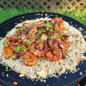 A pile of shrimp fried rice with peas and carrots, eggs, scallion whites and garlic on an open fire skillet on the grill