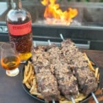 The French Onion Steak Skewers are plated next to a bottle of Pendleton Whisky next to the grill with flames.