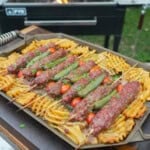 A platter of grilled beef kebabs with waffle fries, cherry tomatoes, sliced white onion and chile peppers