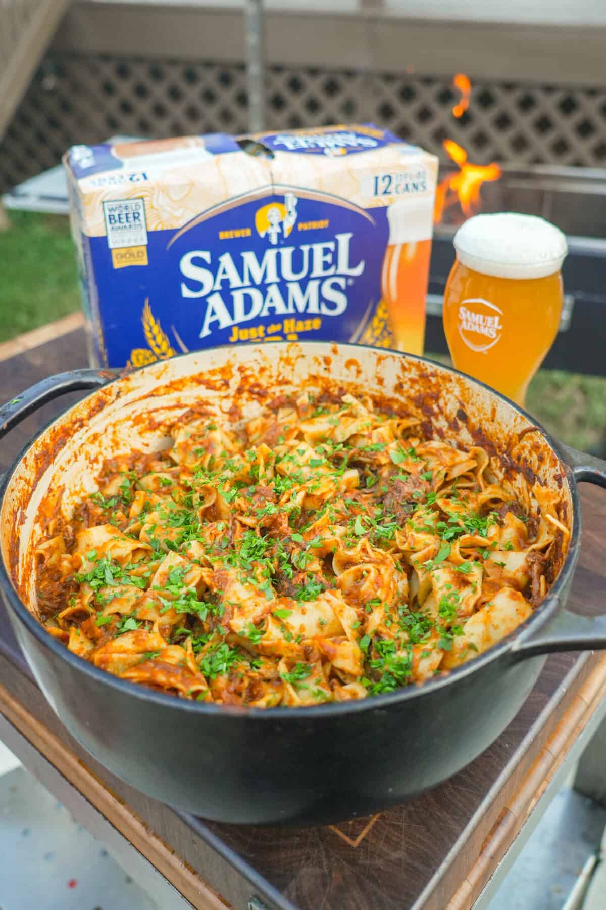 A pot of finished braised beef ragu, garnished with chopped parsley and Parmesan cheese, in front of a pack of Samuel Adams Just the Haze IPA and the beer poured into a glass