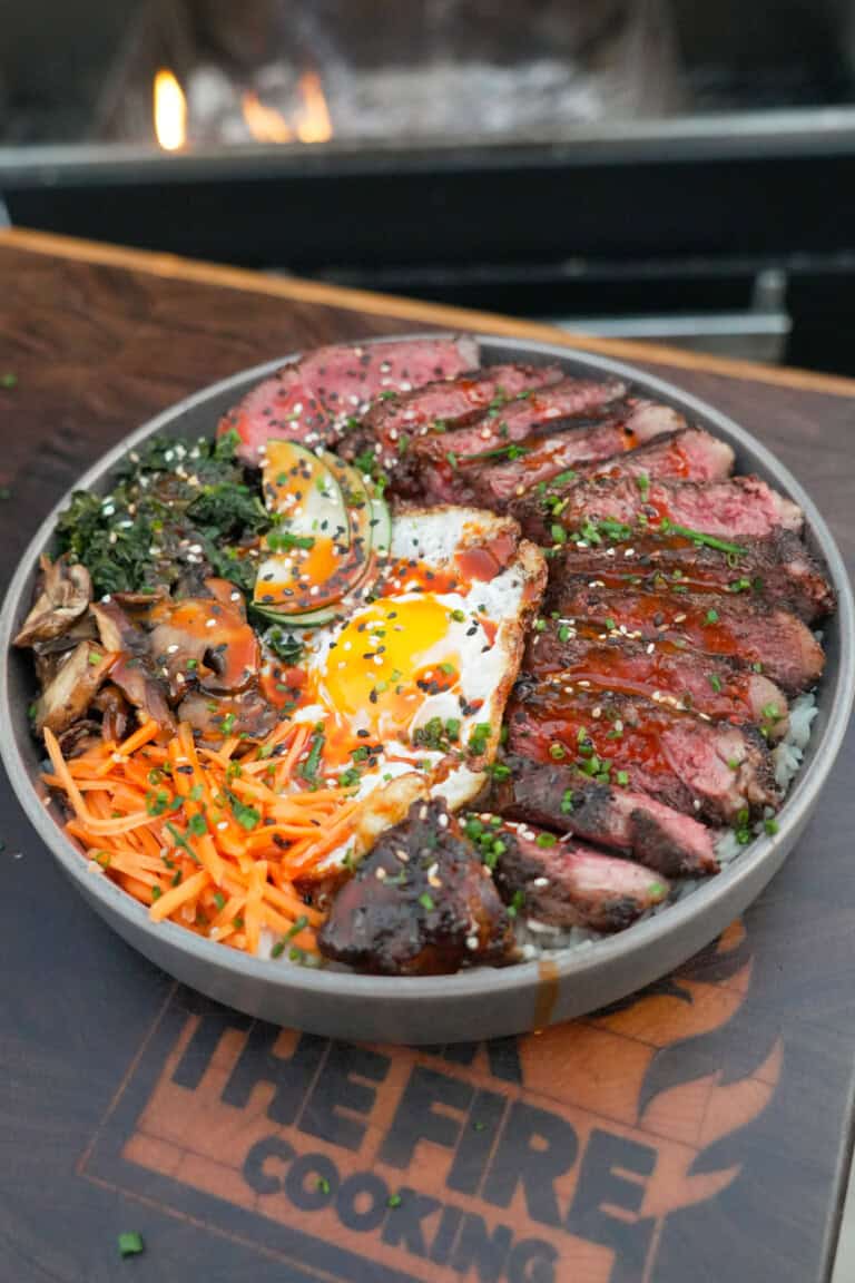 A finished bowl of steak bibimbap, with grilled ribeye, a runny fried egg, bibimbap sauce, carrots, mushrooms, spinach and cucumbers