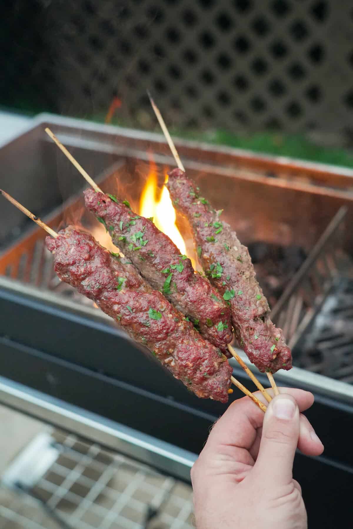 A hand holding three finished grilled beef kebabs on wooden skewers in front of a grill fire