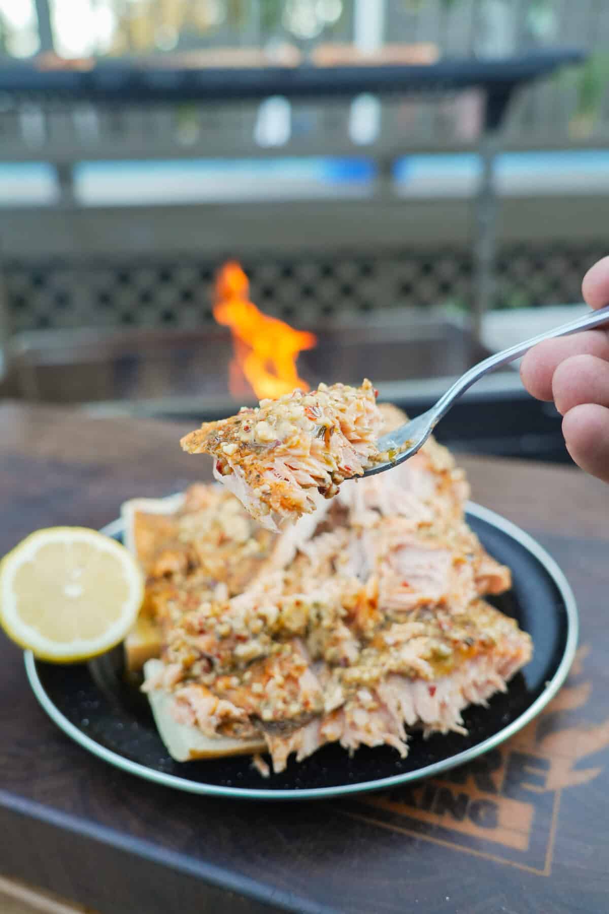 Lifting up a forkful of grilled salmon fillets, with a platter and a lemon half sliced in the background