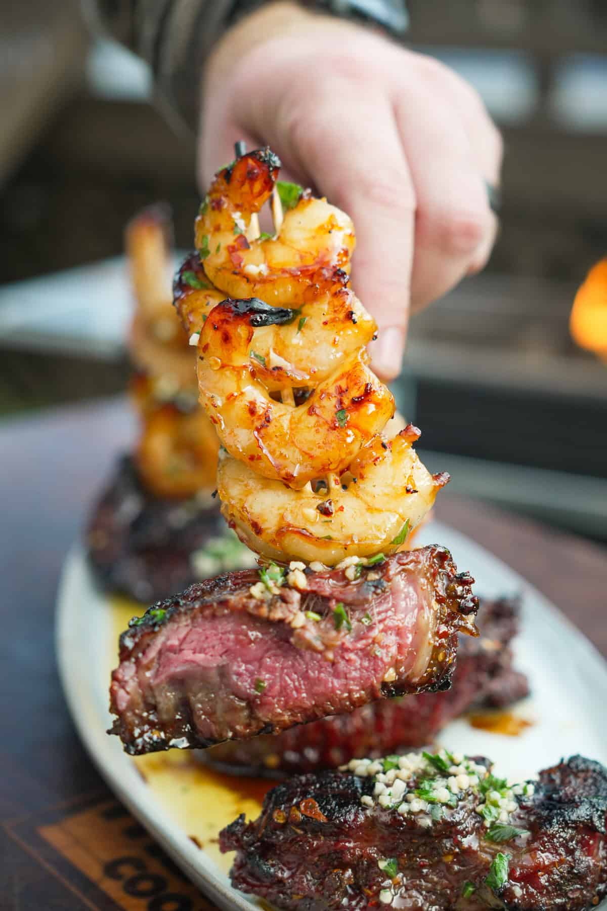 A close up of a hand holding cooked shrimp on a skewer, stacked above slices of filet mignon