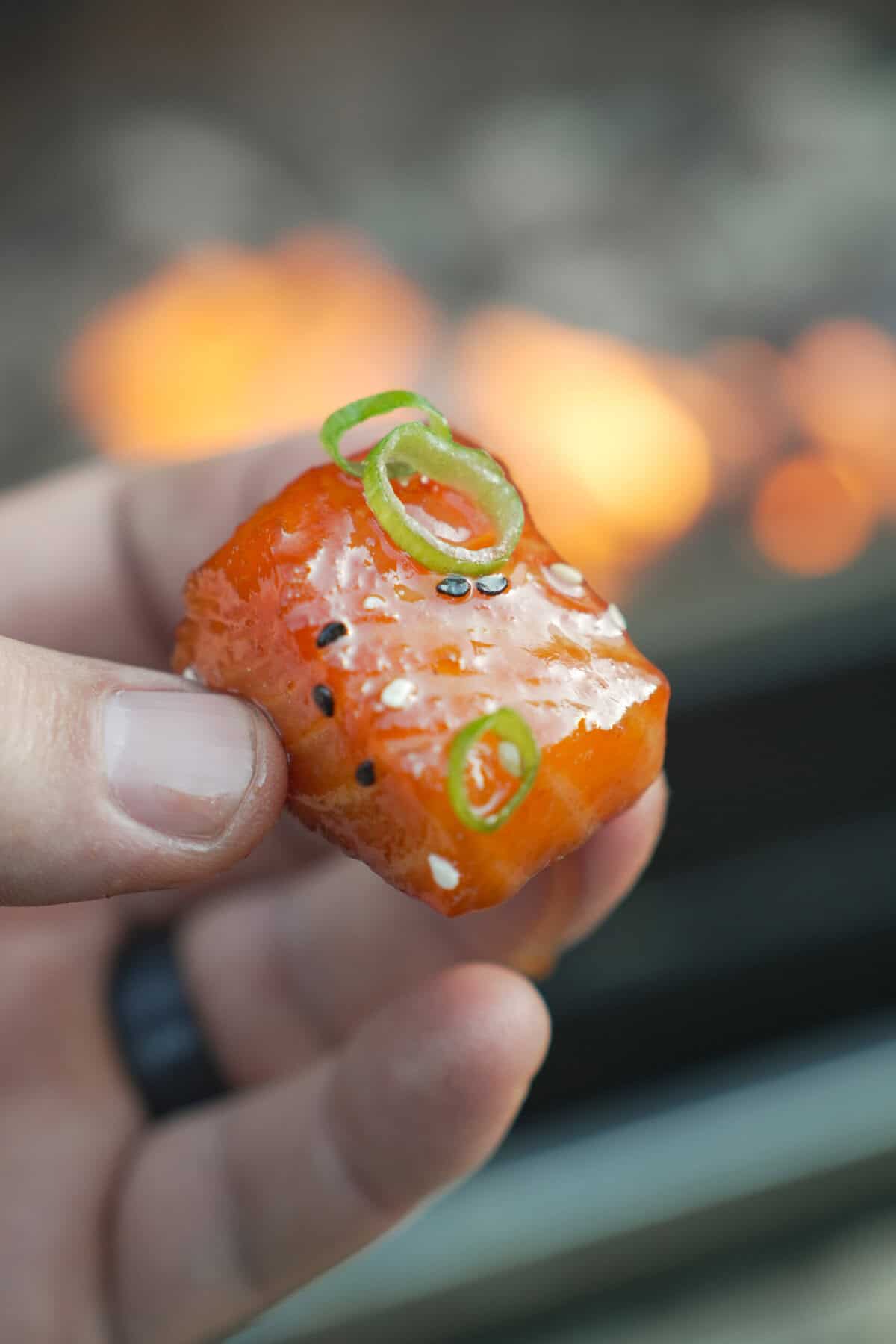 A hand holding a single salmon burnt end for a close up, with the grill fire in the background