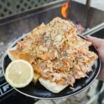 A hand holding a full plate of grilled salmon fillets with cowboy butter, with a sliced lemon half on the side, in front of the FYR Grill