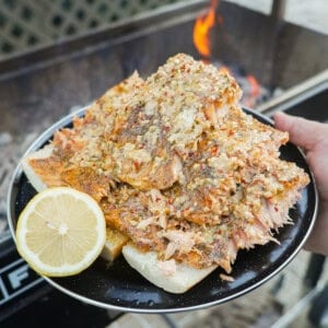 A hand holding a full plate of grilled salmon fillets with cowboy butter, with a sliced lemon half on the side, in front of the FYR Grill