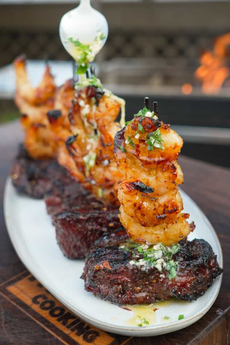 A row of hot honey steak and shrimp on a plate, with filet mignon as the base and shrimp skewers stacked on top of the steak
