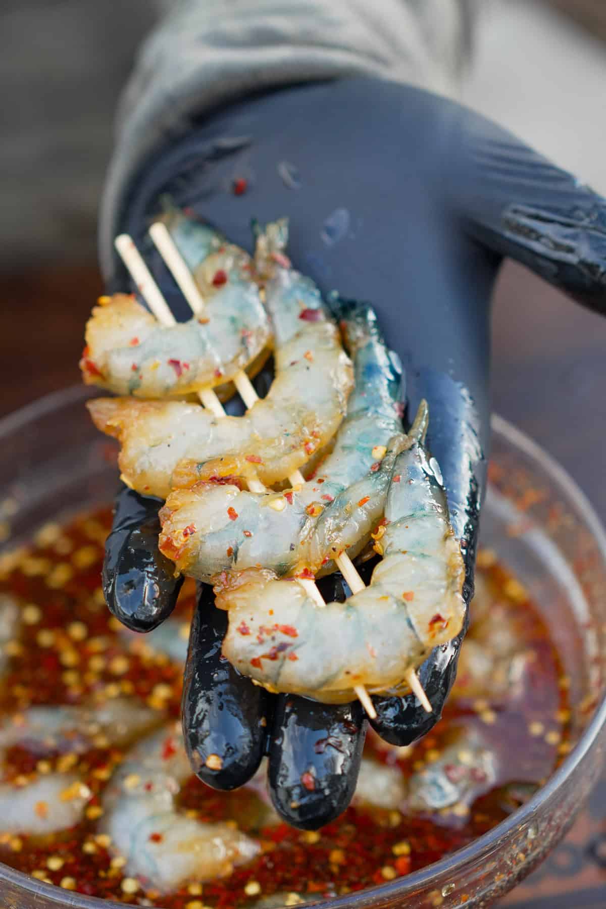 A gloved hand holding raw skewered shrimp above a bowl of hot honey cure
