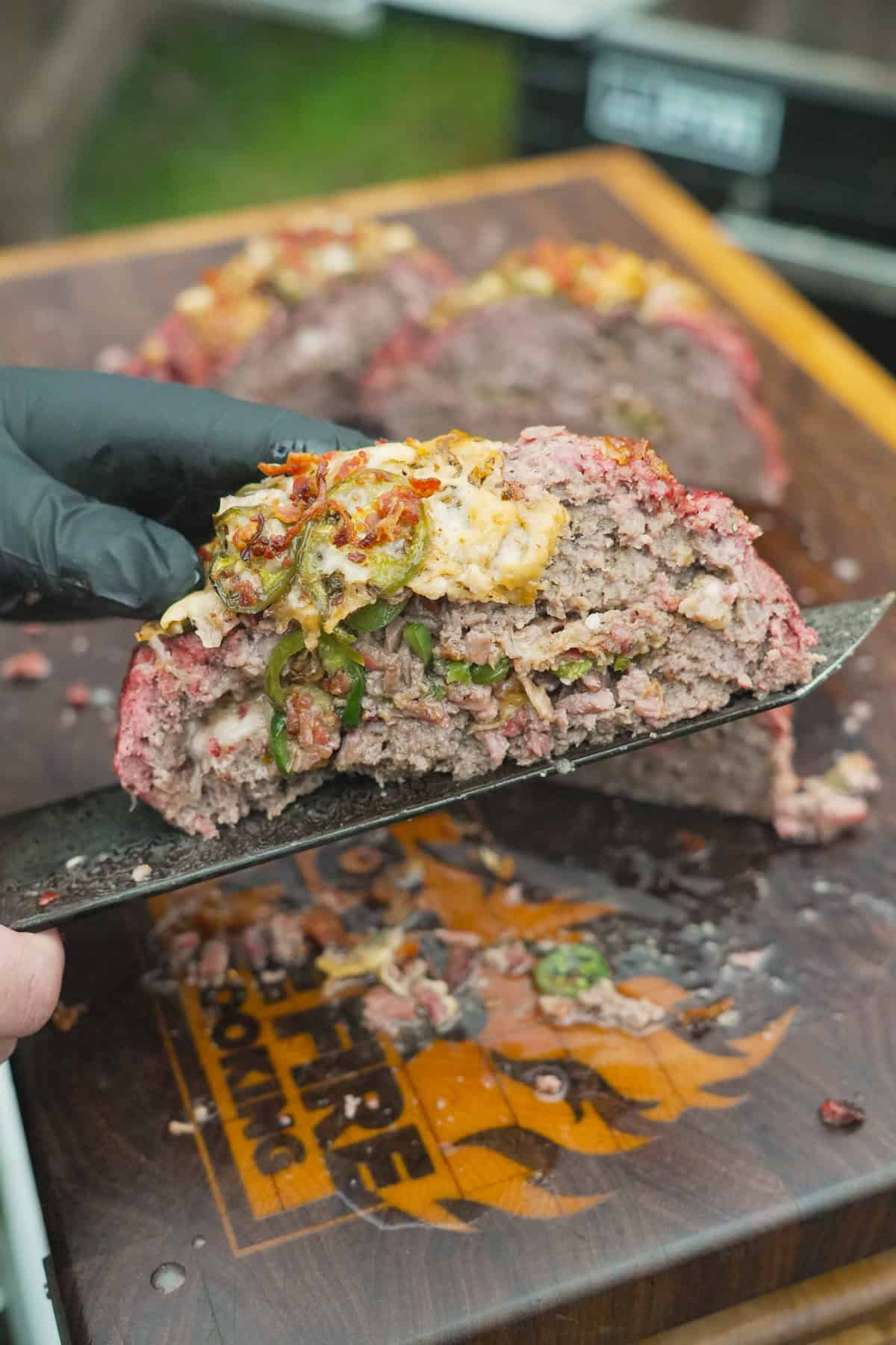 A gloved hand holding up a slice of Texas Twinkie meatloaf balanced on a chef's knife, with cheese, brisket, jalape&ntilde;os and bacon bits on the inside