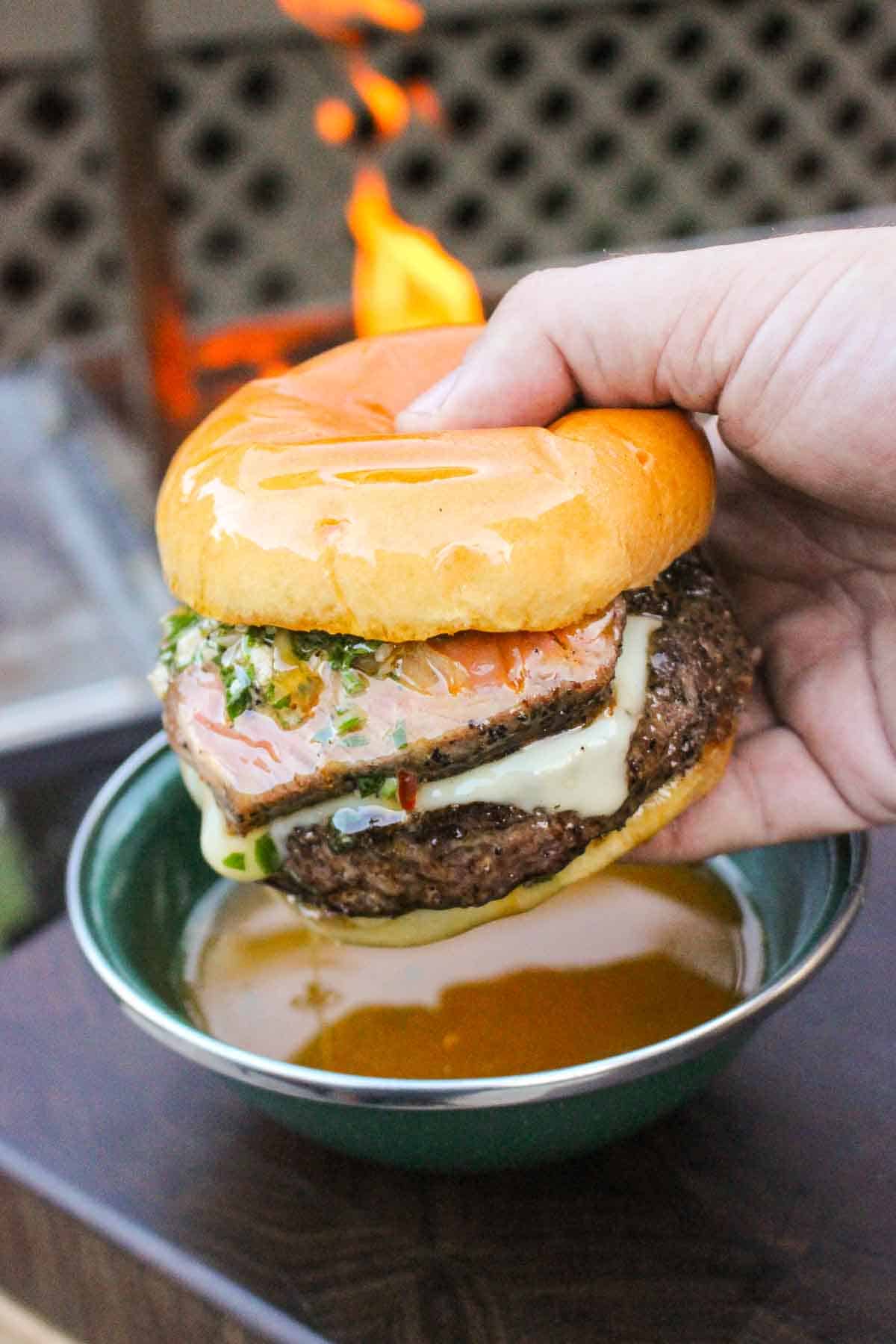 A hand holding a burger over a bowl of cowboy butter for dipping with a live grill fire in the background