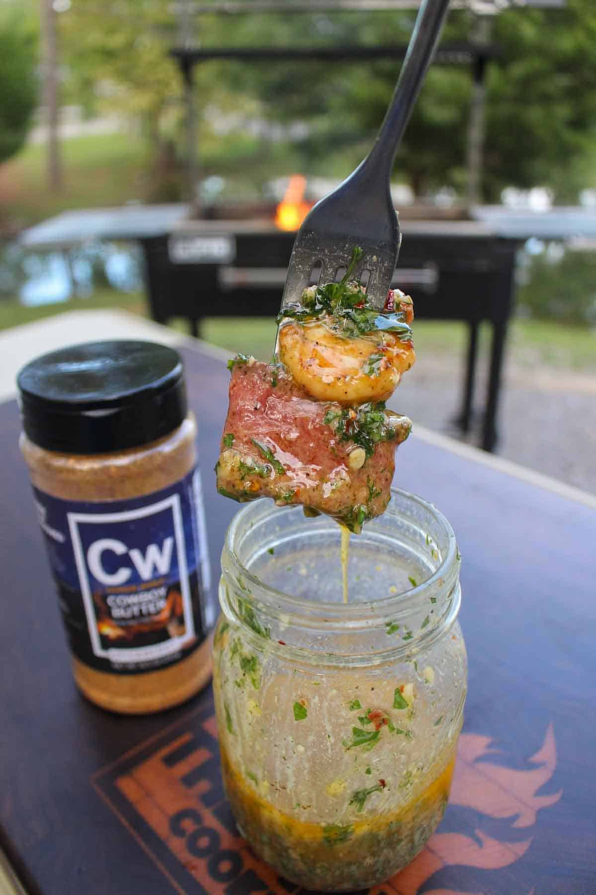 A forkful of steak and shrimp, held above a mason jar filled with cowboy butter and Cowboy Butter Seasoning jar in the background