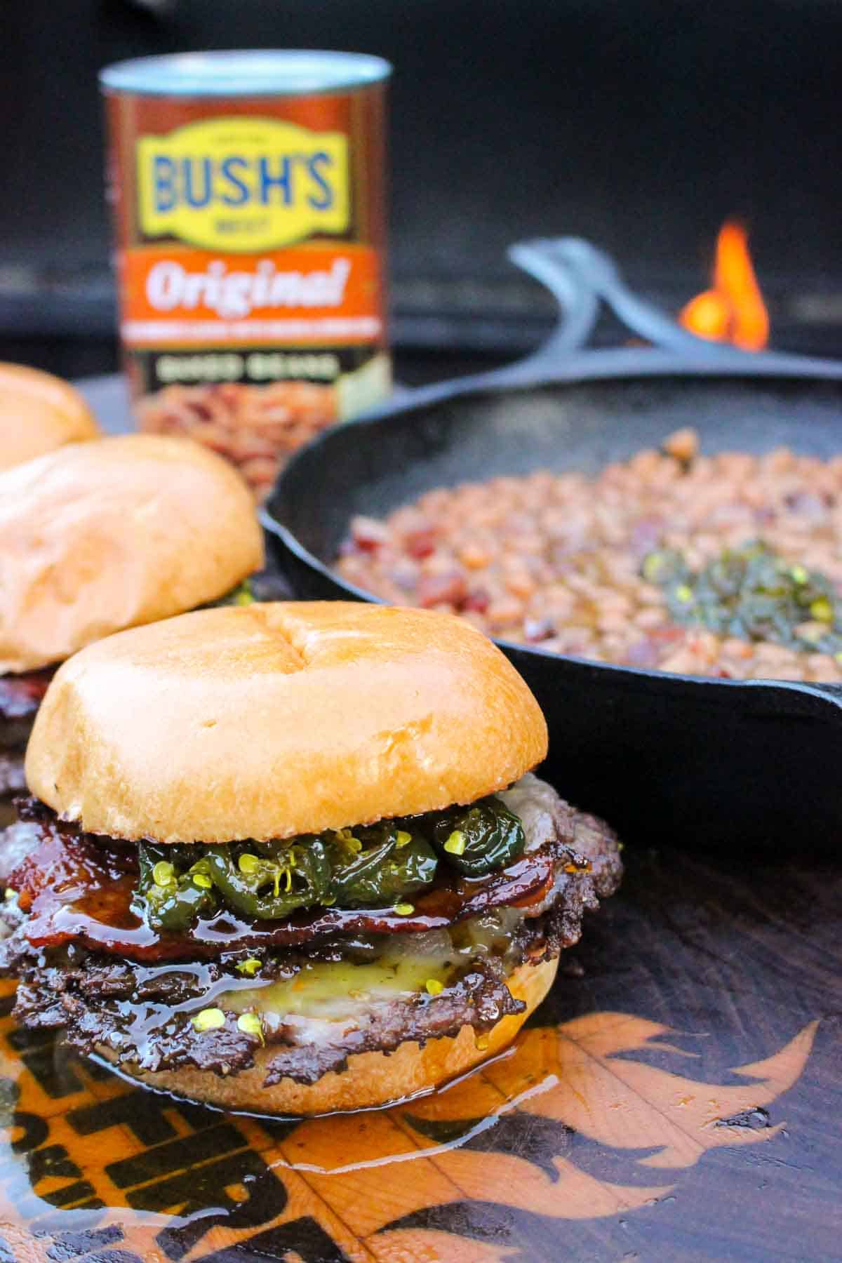 A lineup of cowboy candy burgers with baked beans in a cast iron skillet in the background for cowboy recipes