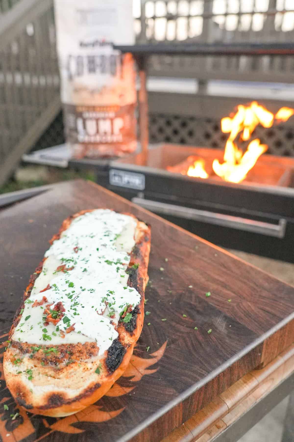 A finished loaf of chicken bacon ranch stuffed bread sitting on a cutting board, with a bag of Cowboy Charcoal and a grill fire in the background