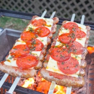 Two rows of hot honey pizza sausage skewers on the grill, with the sausages threaded on two two-pronged skewers and topped with marinara sauce, mozzarella cheese, pepperoni slices and fresh basil