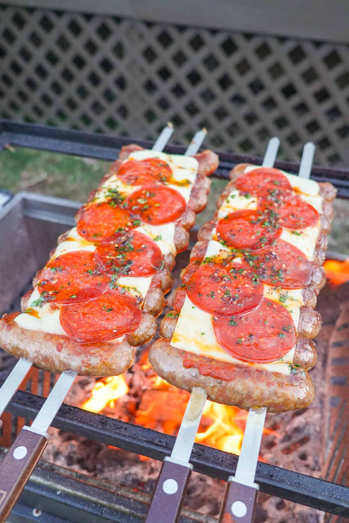 Two rows of hot honey pizza sausage skewers on the grill, with the sausages threaded on two two-pronged skewers and topped with marinara sauce, mozzarella cheese, pepperoni slices and fresh basil