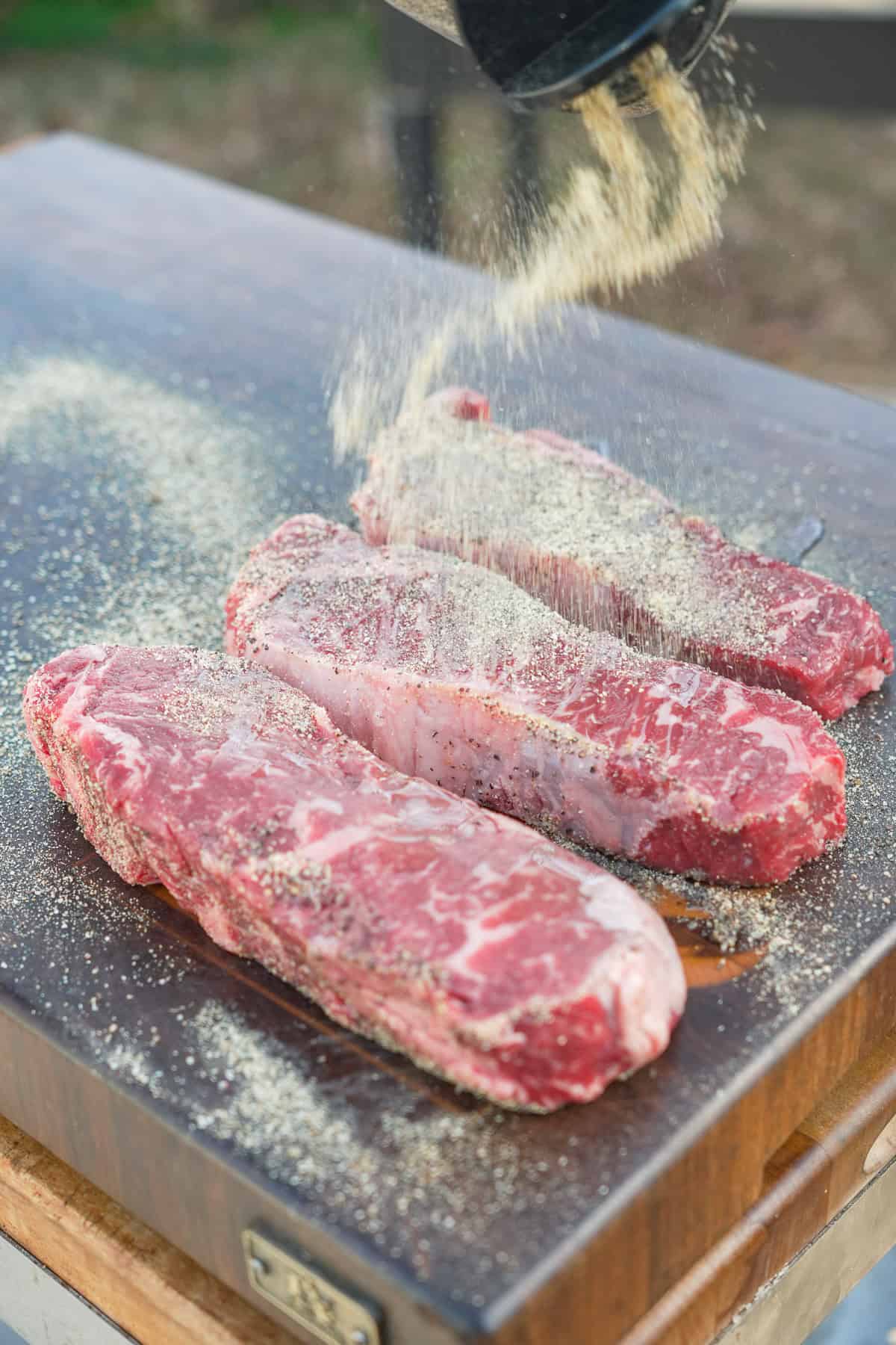 Seasoning three raw steaks on a cutting board with Salt Pepper Garlic Seasoning