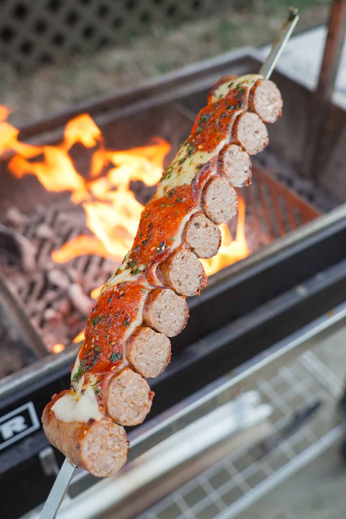 Sliced sausage skewers in front of the grill fire, cut in half to reveal the inside of the and the pizza ingredients on top