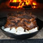 A pile of grilled beef shawarma on a bed of onions in front of a roaring pizza oven fire