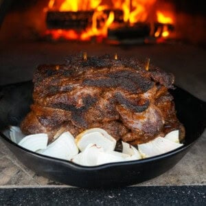 A pile of grilled beef shawarma on a bed of onions in front of a roaring pizza oven fire