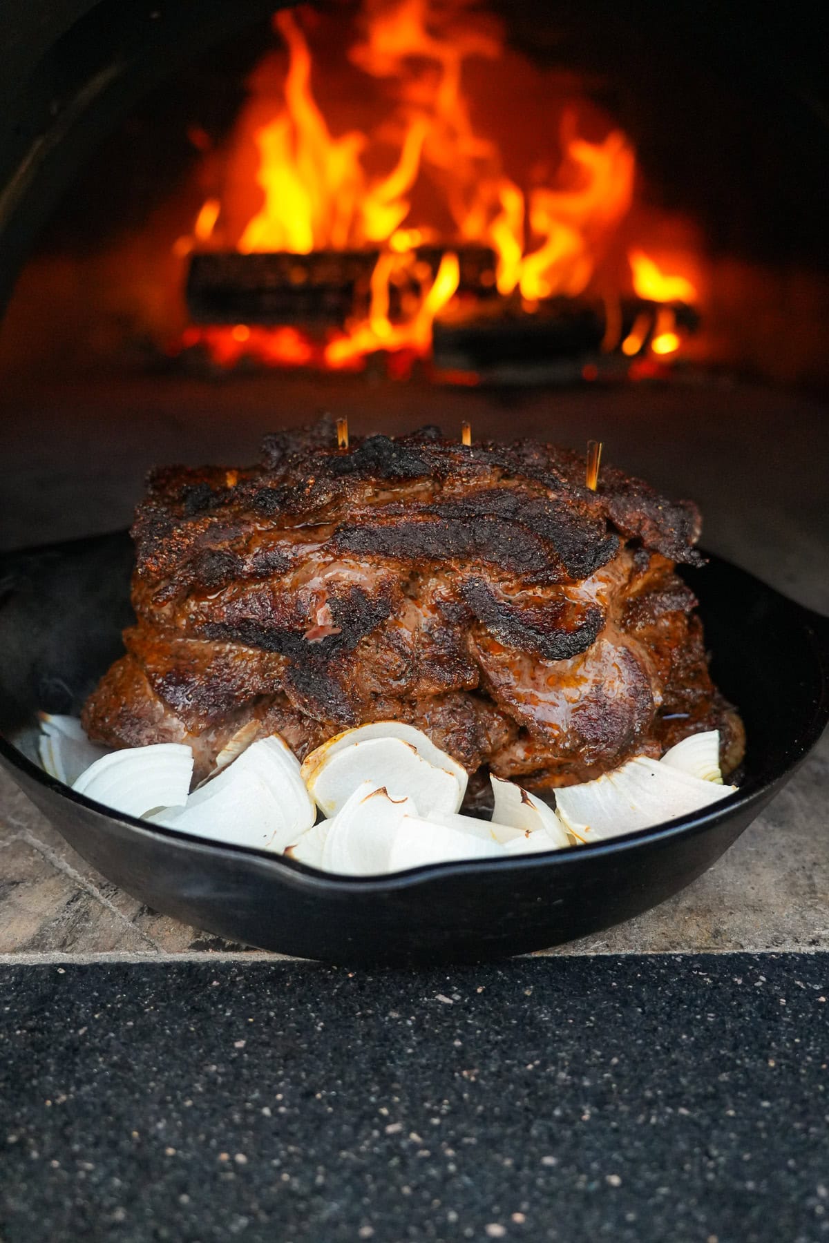 A pile of grilled beef shawarma on a bed of onions in front of a roaring pizza oven fire