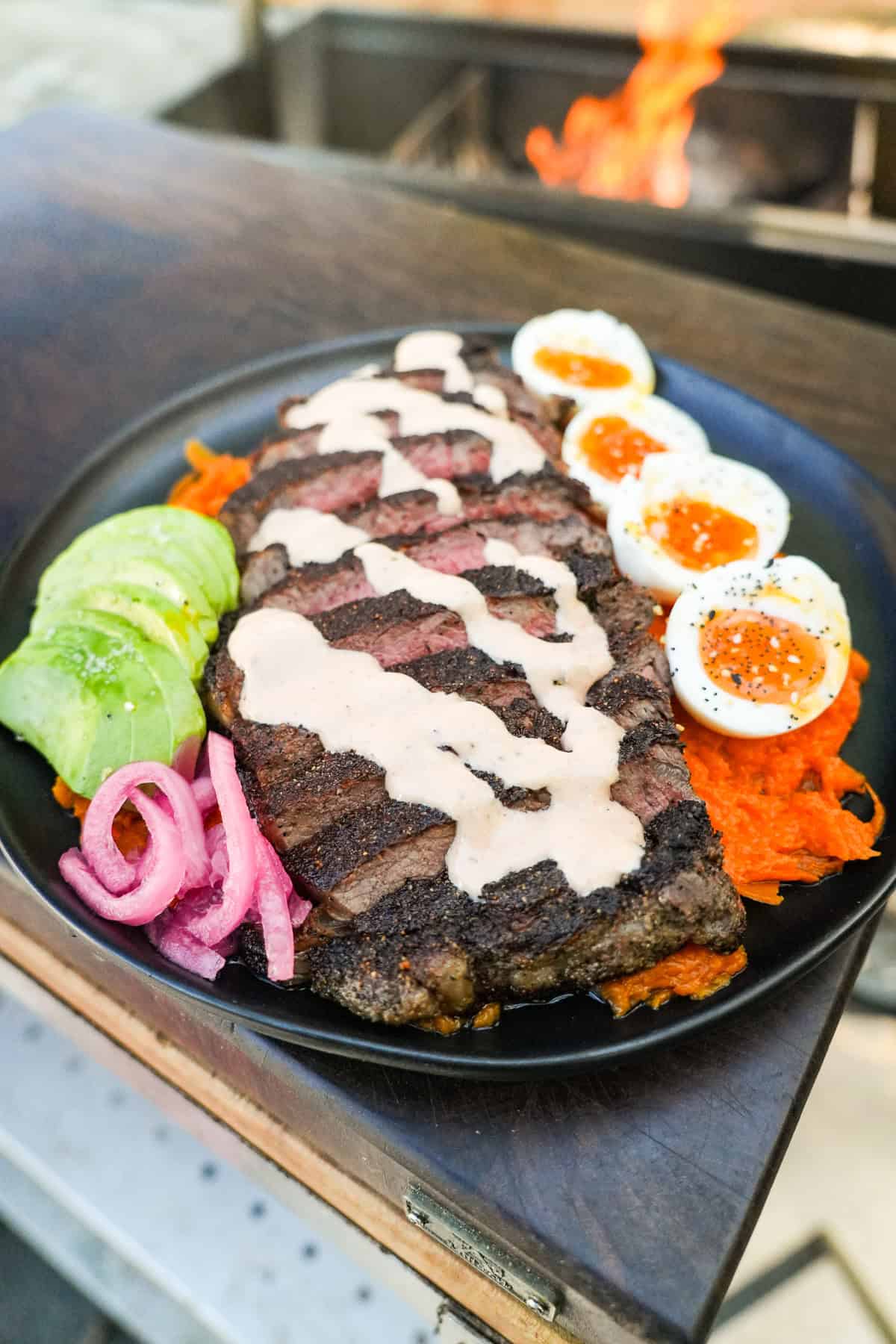 A finished healthy steak bowl in front of the grill fire, with sliced steak, honey chipotle sauce, soft boiled eggs, avocado, pickled red onions and mashed sweet potato