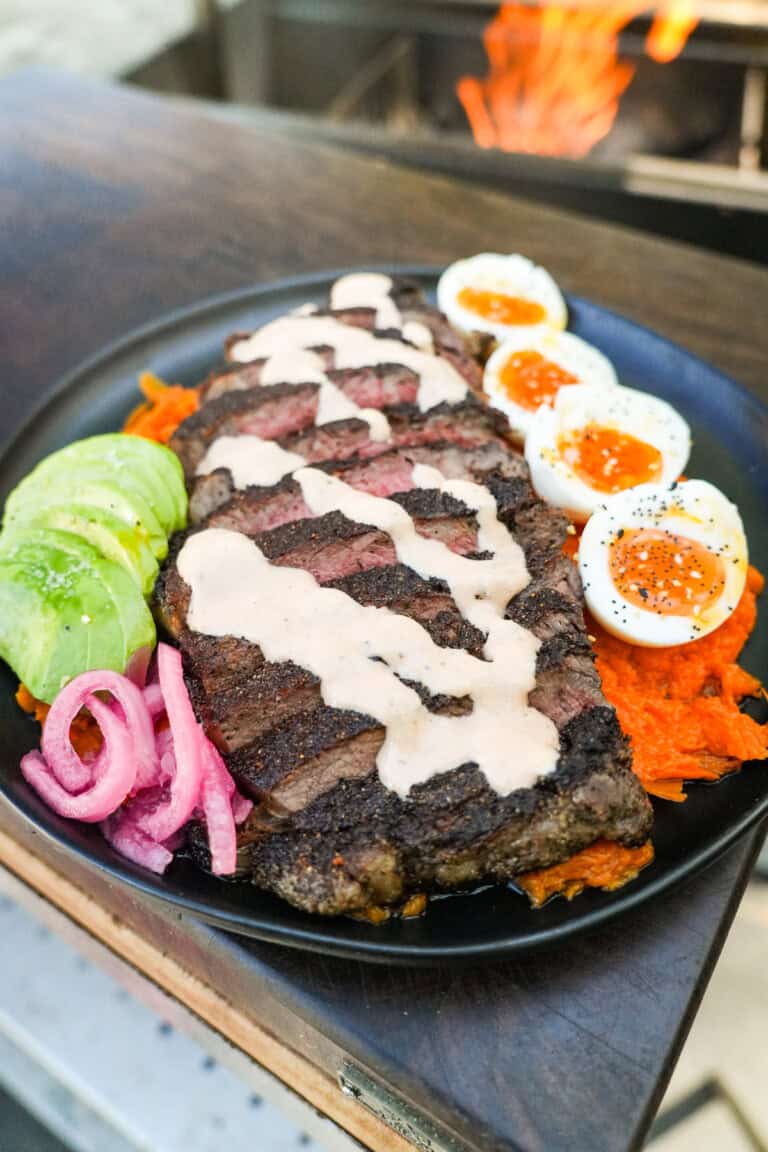 A finished healthy steak bowl with sliced steak, honey chipotle sauce, soft boiled eggs, avocado, pickled red onions and mashed sweet potato