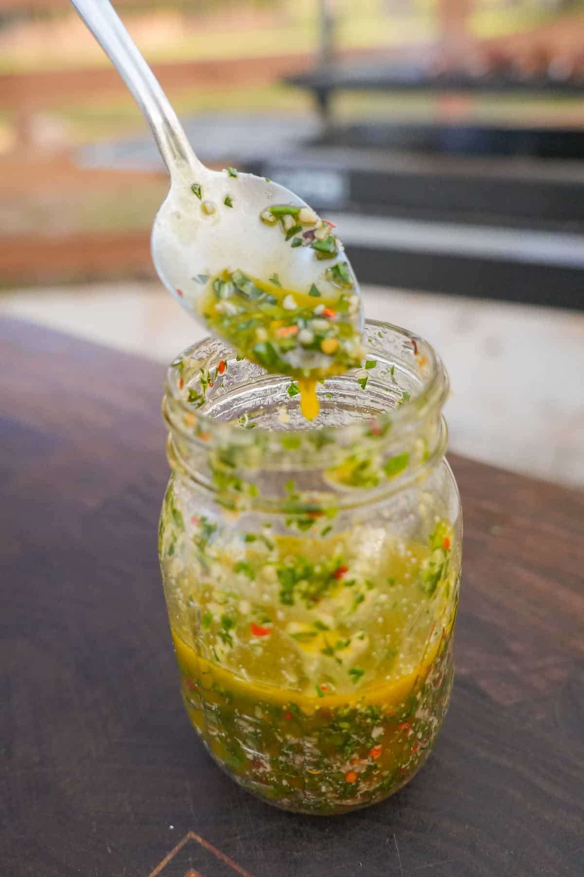 A spoon above a jar of Italian chimichurri sauce, with basil, parsley, Parmesan, garlic, olive oil, red wine vinegar and red chili flakes