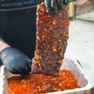 Derek Wolf wearing gloves and holding a rack of spare ribs above a foil pan filled with bourbon peach glaze