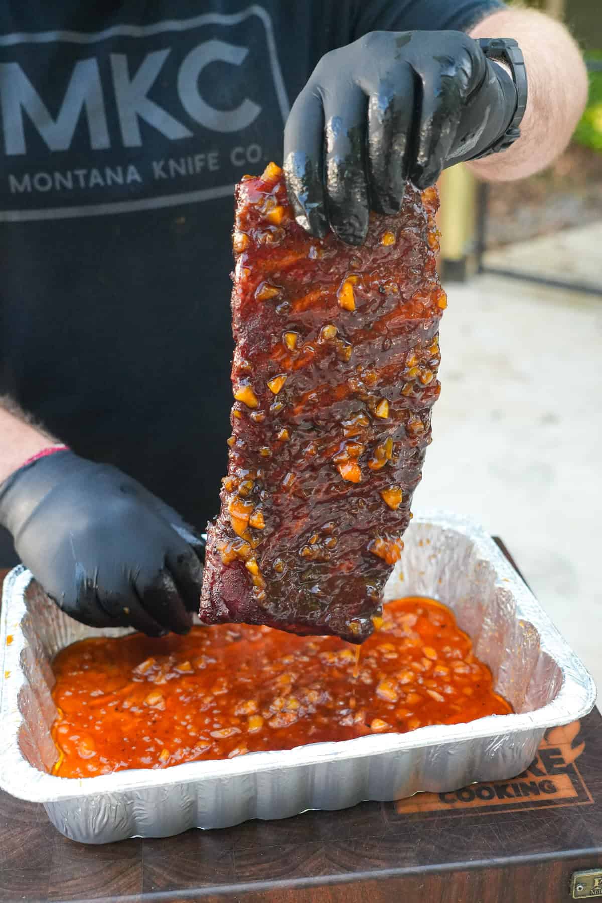 Derek Wolf wearing gloves and holding a rack of spare ribs above a foil pan filled with bourbon peach glaze