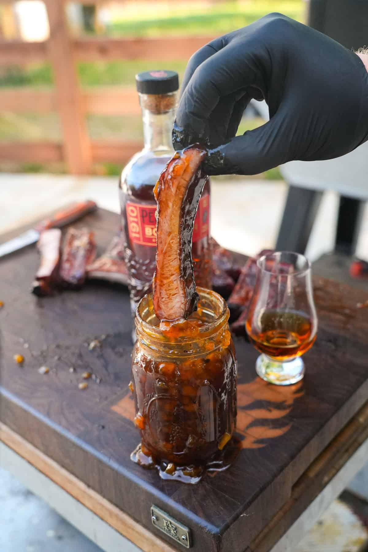 Derek Wolf dipping a single rib in the bourbon peach glaze in a mason jar with Pendleton Bourbon in the background