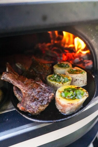 A cast iron skillet with lamb chops, bone marrow rounds filled with mint chimichurri in a pizza oven, with a live fire in the background