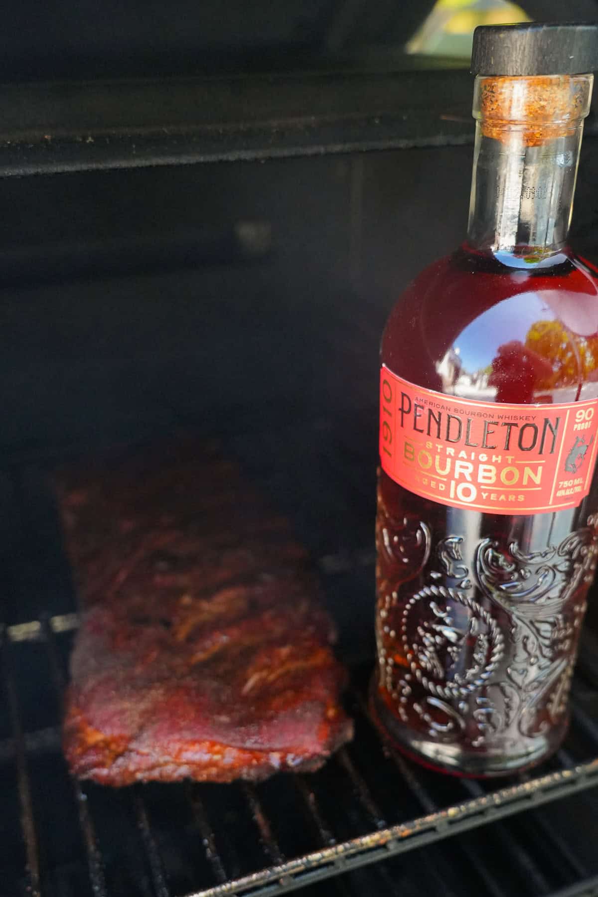 A rack of ribs on the smoker with a bottle of Pendleton 1910 Bourbon in the foreground