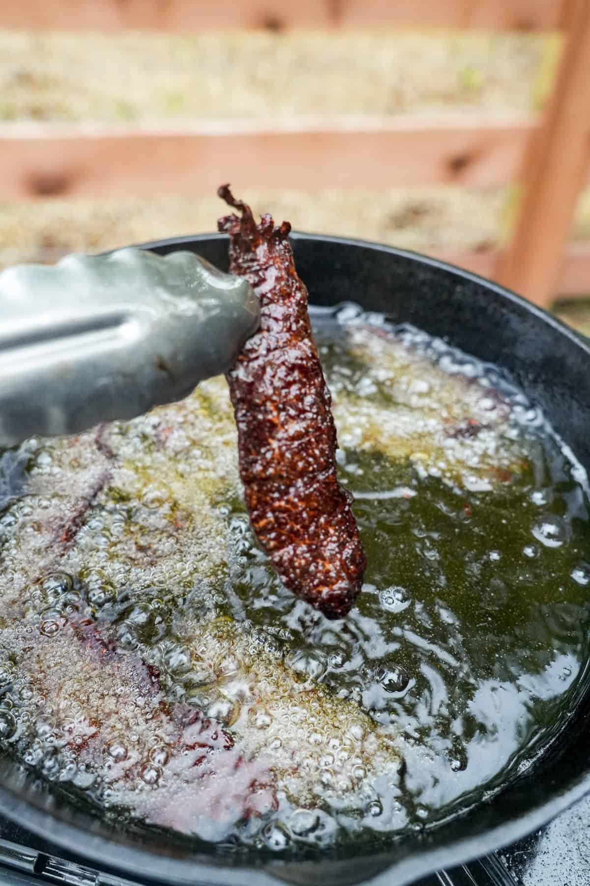 Using tongs to pull a slice of crispy brisket jerky out of a vat of oil after frying